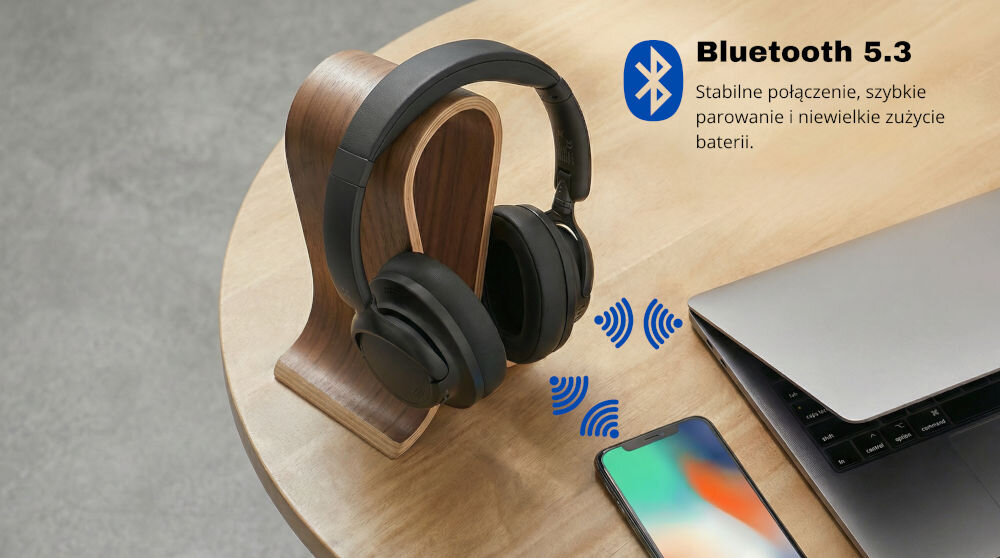 Na drewnianym stojaku znajdują się czarne słuchawki nauszne. Obok widoczny jest telefon, laptop oraz tekst: \'Bluetooth 5.3 Stabilne połączenie, szybkie parowanie i niewielkie zużycie baterii\'.