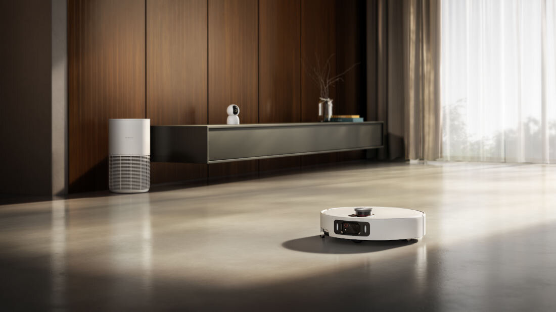 Na podłodze w nowoczesnym wnętrzu znajduje się biały robot sprzątający. W tle widać drewnianą ścianę, półkę z książkami i dekoracjami oraz duże okno z zasłonami.