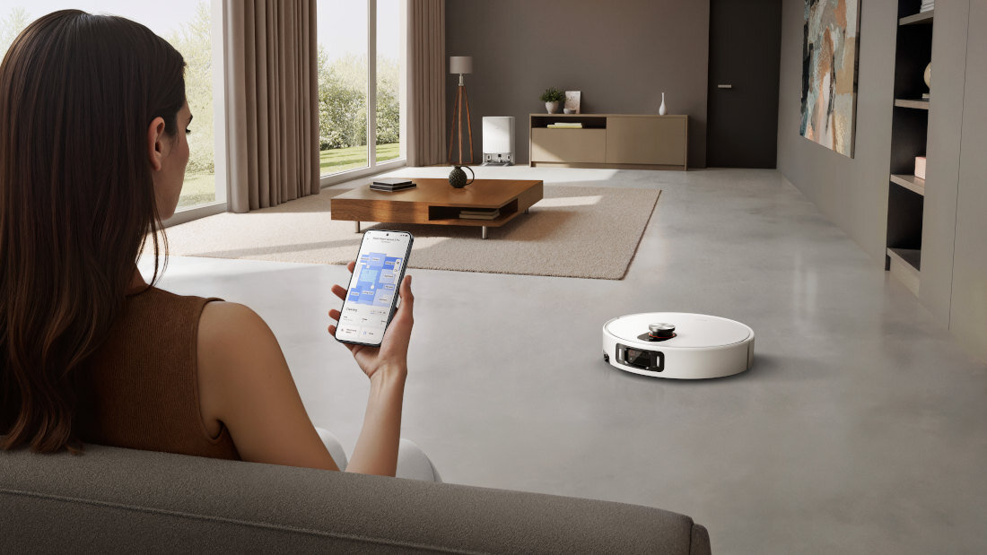 Osoba siedzi na kanapie, trzymając w ręku smartfon z aplikacją. Na podłodze w nowoczesnym salonie znajduje się biały robot sprzątający.