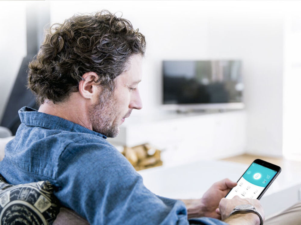 Mężczyzna siedzi na kanapie, trzymając w ręku smartfon z widocznym ekranem. W tle znajduje się telewizor na białej szafce.