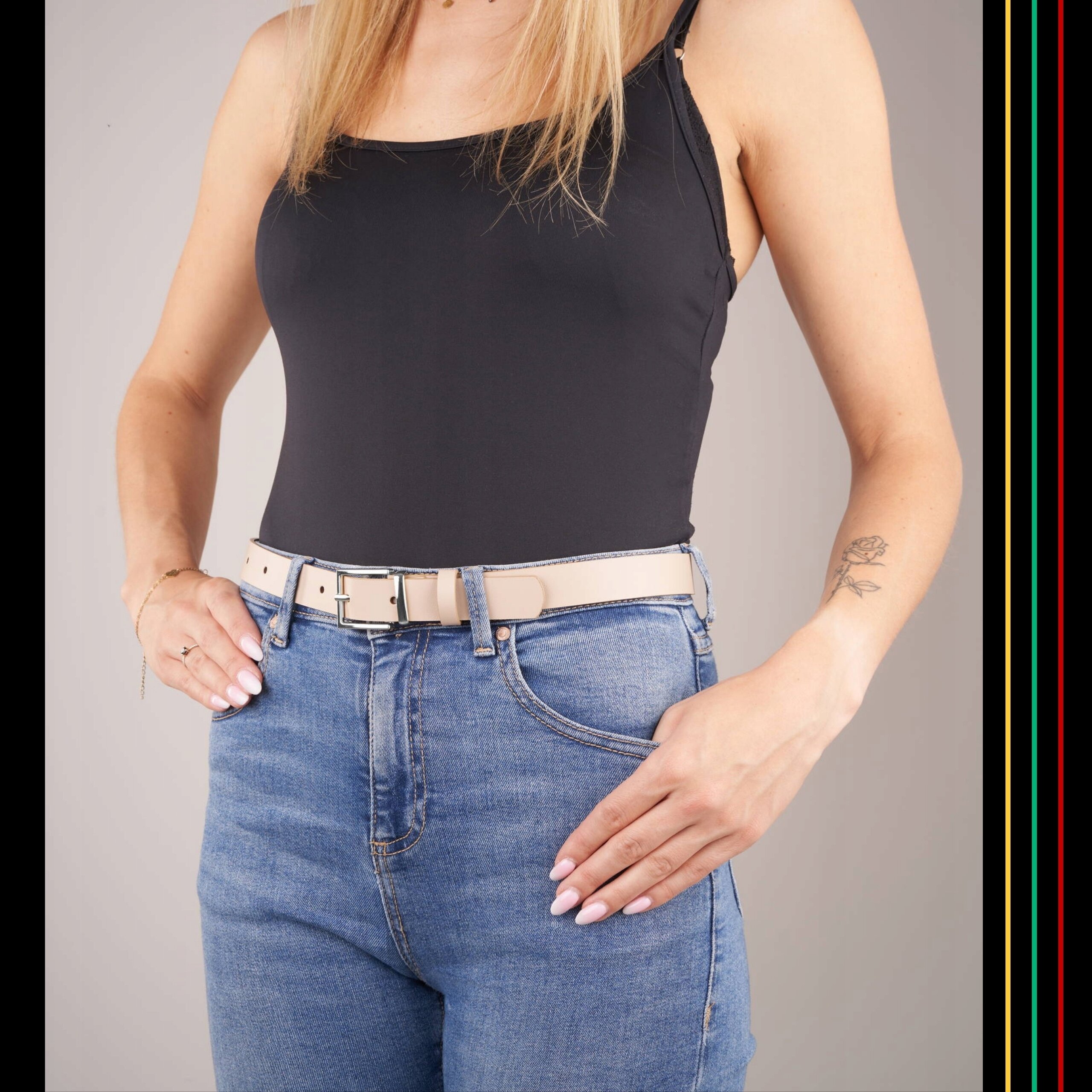 Osoba ubrana w czarną bluzkę i niebieskie dżinsy prezentuje beżowy skórzany pasek z srebrną klamrą. Na ręce widoczny jest tatuaż w kształcie róży.
