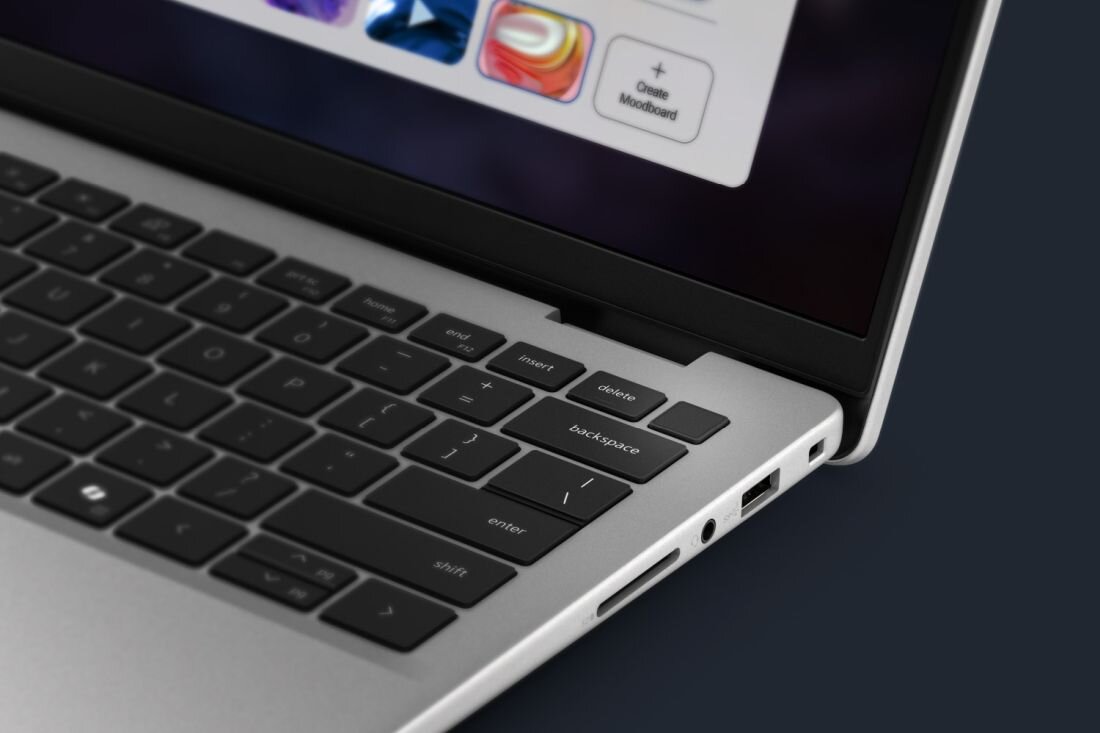 Zbliżenie na klawiaturę laptopa z widocznymi klawiszami, takimi jak \'enter\', \'shift\', \'delete\', oraz portami bocznymi. Na ekranie widać fragment interfejsu z kolorowymi ikonami i przyciskiem \'+ Create Moodboard\'.