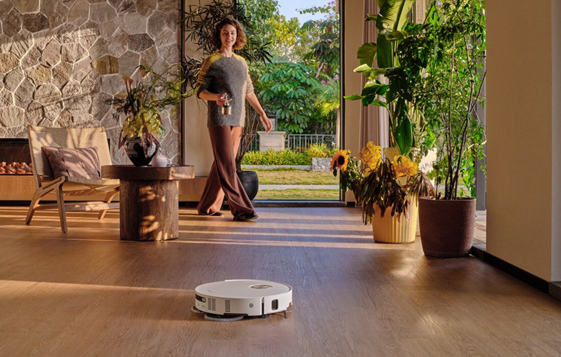 Na drewnianej podłodze znajduje się biały robot sprzątający. W tle widać osobę stojącą w jasnym pomieszczeniu z dużymi roślinami i widokiem na ogród.