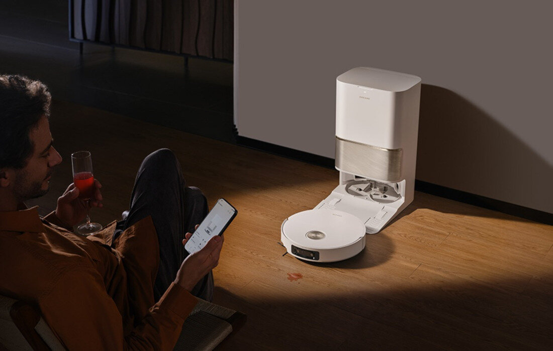 Na drewnianej podłodze znajduje się biały robot sprzątający z otwartą stacją dokującą. Obok siedzi osoba trzymająca smartfon i szklankę z czerwonym napojem.