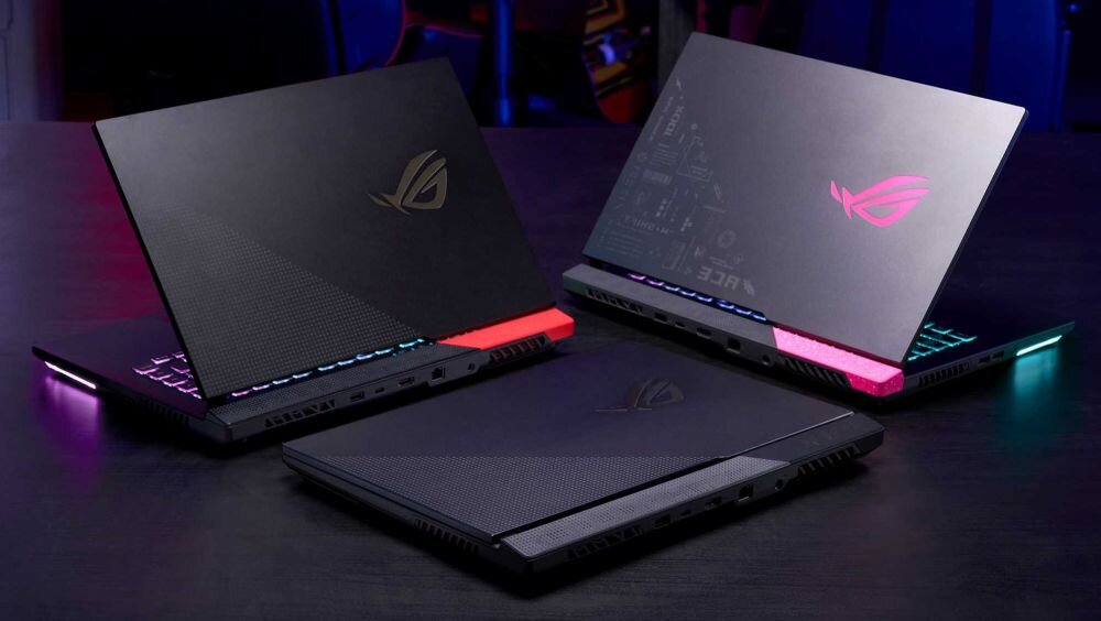 Trzy laptopy z podświetlanymi klawiaturami i logo ROG na obudowach, ustawione na ciemnym stole. Każdy z laptopów ma różnokolorowe podświetlenie na tylnej krawędzi.