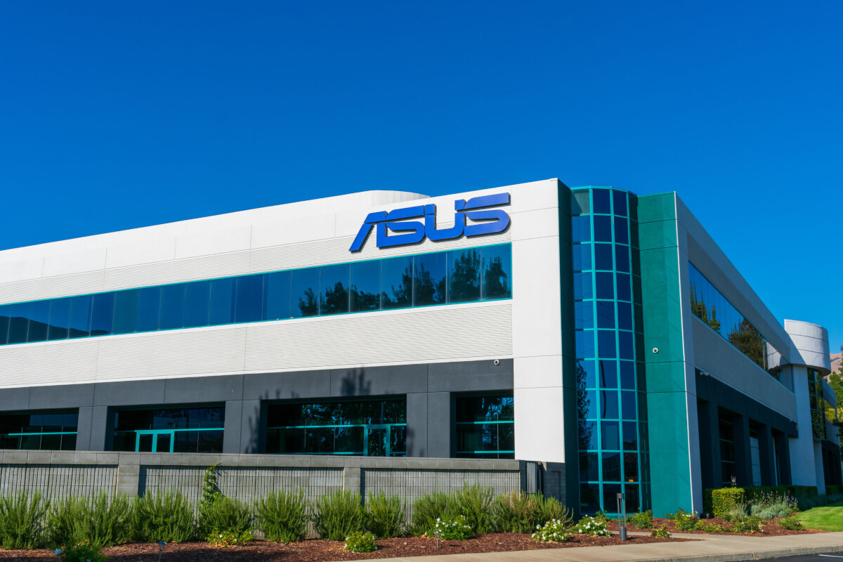 Budynek z dużym logo \'ASUS\' na białej fasadzie, z przeszklonymi oknami i zielonymi elementami architektonicznymi. Przed budynkiem znajduje się zadbana zieleń.