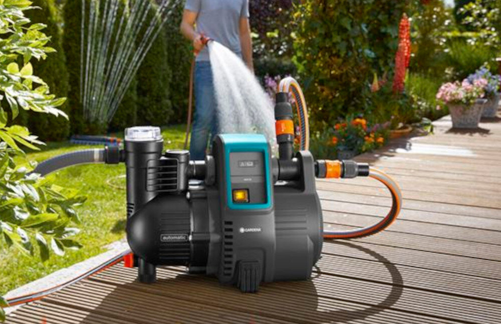 Na drewnianym tarasie znajduje się elektryczny hydrofor z widocznym logo GARDENA. W tle osoba podlewa ogród za pomocą węża ogrodowego.