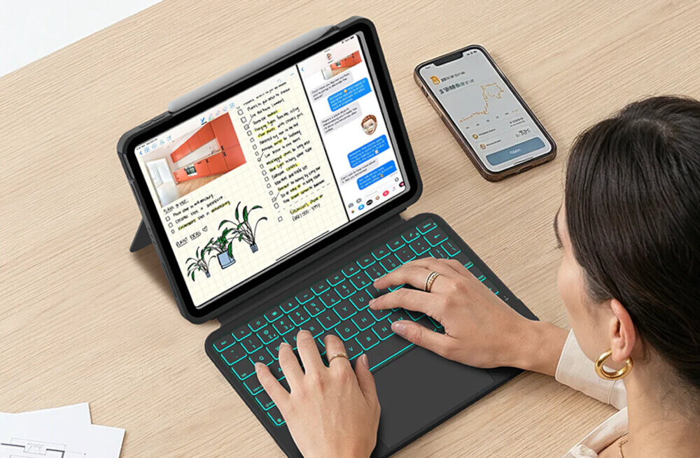 Osoba korzysta z tabletu w etui z klawiaturą, na ekranie widoczne są notatki i wiadomości. Obok leży smartfon z wykresem na ekranie.