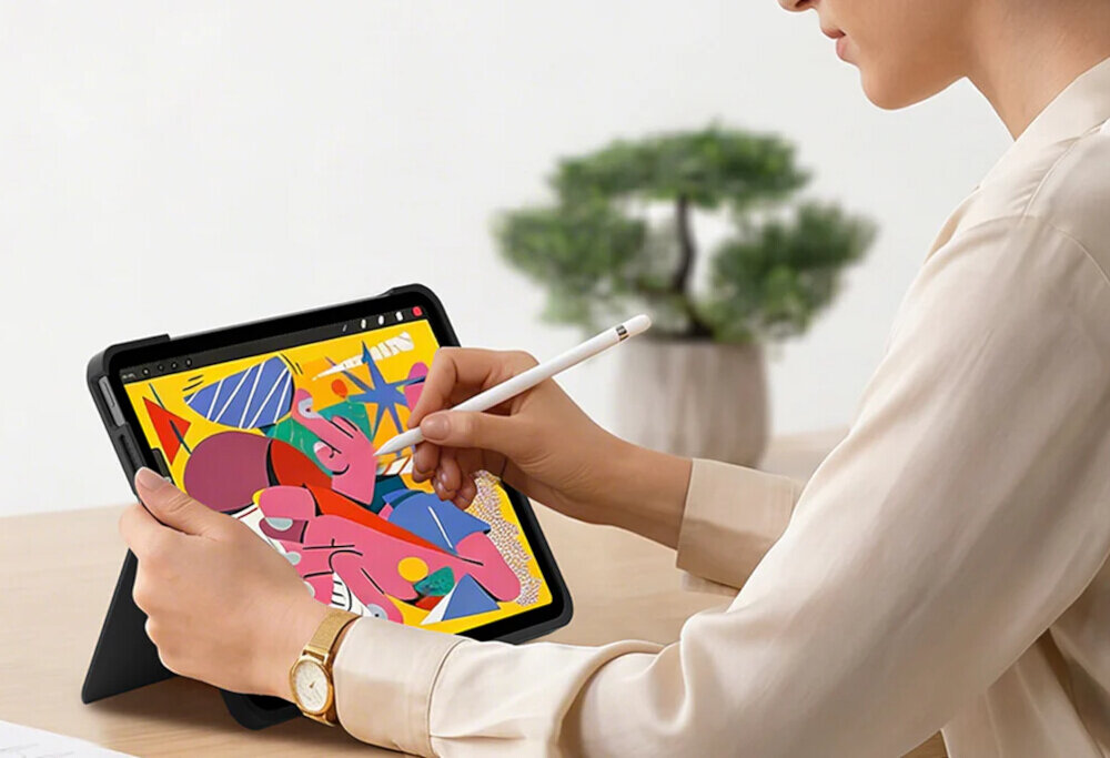 Osoba trzyma tablet z kolorową grafiką na ekranie, używając rysika. Tablet jest umieszczony w czarnym etui, a w tle widać rozmytą roślinę.