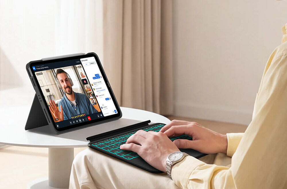 Osoba siedzi przy stoliku, używając czarnej klawiatury z podświetleniem, połączonej z tabletem w etui. Na ekranie tabletu widoczna jest wideorozmowa z mężczyzną oraz aplikacja do czatu.