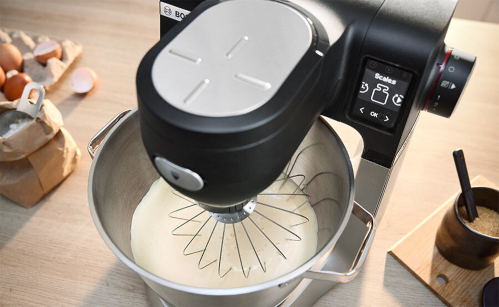 Robot kuchenny z misą, w której znajduje się mieszana masa. W tle widoczne są jajka, mąka i cukier w pojemniku.