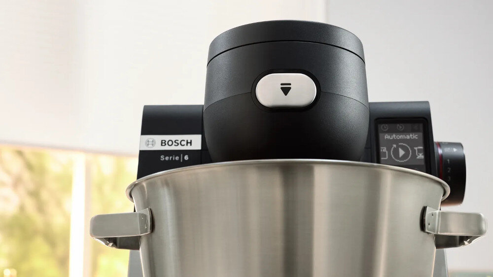 Na zdjęciu widoczny jest fragment robota kuchennego z metalową misą i czarną obudową. Na obudowie znajduje się logo \'BOSCH Serie | 6\'.