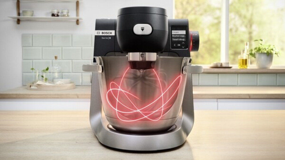 Robot kuchenny z logo BOSCH na blacie kuchennym, z widocznym metalowym mikserem i czerwonymi liniami świetlnymi w misie. W tle jasna kuchnia z oknem i półkami.