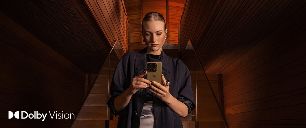 Osoba trzyma smartfon w drewnianym wnętrzu. W lewym dolnym rogu widoczne jest logo \'Dolby Vision\'.