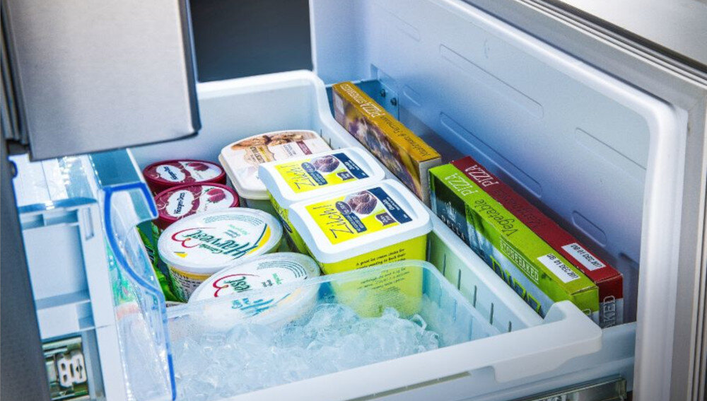 Otwarta szuflada zamrażarki z różnymi produktami spożywczymi, w tym pudełkami lodów i opakowaniami pizzy. W szufladzie widoczne są również kostki lodu.