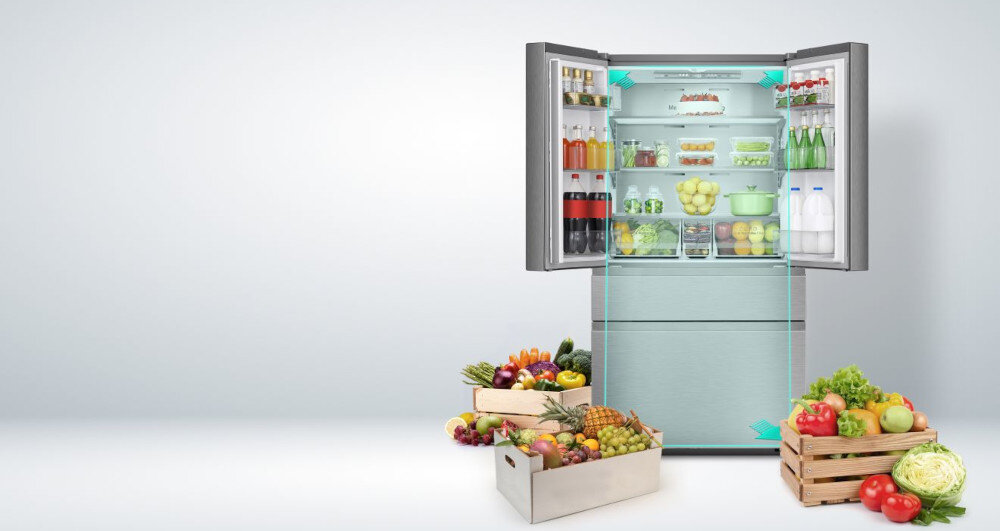 Otwarta lodówka z widocznymi półkami wypełnionymi różnorodnymi produktami spożywczymi, takimi jak owoce, warzywa, napoje i nabiał. Przed lodówką znajdują się skrzynki z owocami i warzywami.