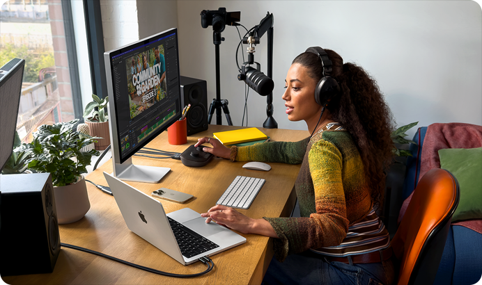 Osoba siedzi przy biurku z laptopem marki APPLE, obok którego znajduje się monitor z otwartym programem do edycji wideo. Na biurku leżą także klawiatura, myszka, notatnik i roślina w doniczce.