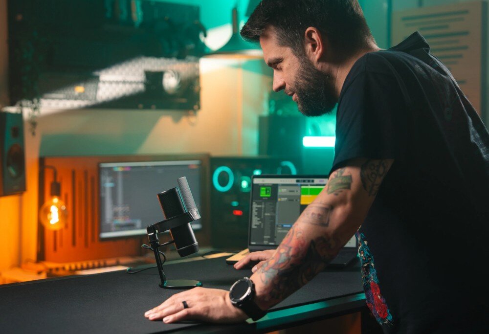 Mikrofon ENDORFY Solum 2 Pro Mężczyzna opiera się o biurko i patrzy na stojący mikrofon pojemnościowy w koszu antywstrząsowym w otoczeniu sprzętu komputerowego