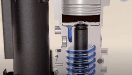 Obraz przedstawia przekrój mechanizmu z widocznymi elementami metalowymi i sprężyną w kolorze niebieskim. W tle widoczna jest część obudowy w kolorze czarnym.