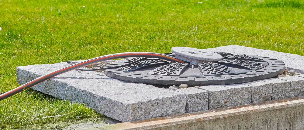 Na obrazie widoczna jest pokrywa studzienki z wężem ogrodowym na tle trawnika. Pokrywa wykonana jest z ciemnego materiału i ma wzór kratki.