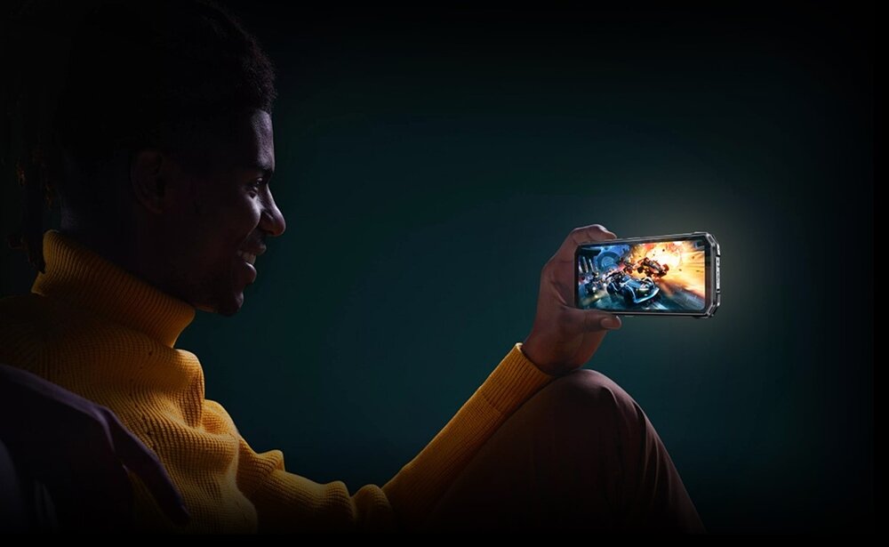 Osoba w żółtym swetrze trzyma smartfon, na którym wyświetlana jest dynamiczna gra z motywem wyścigów i eksplozji. Tło jest ciemne, co podkreśla ekran urządzenia.