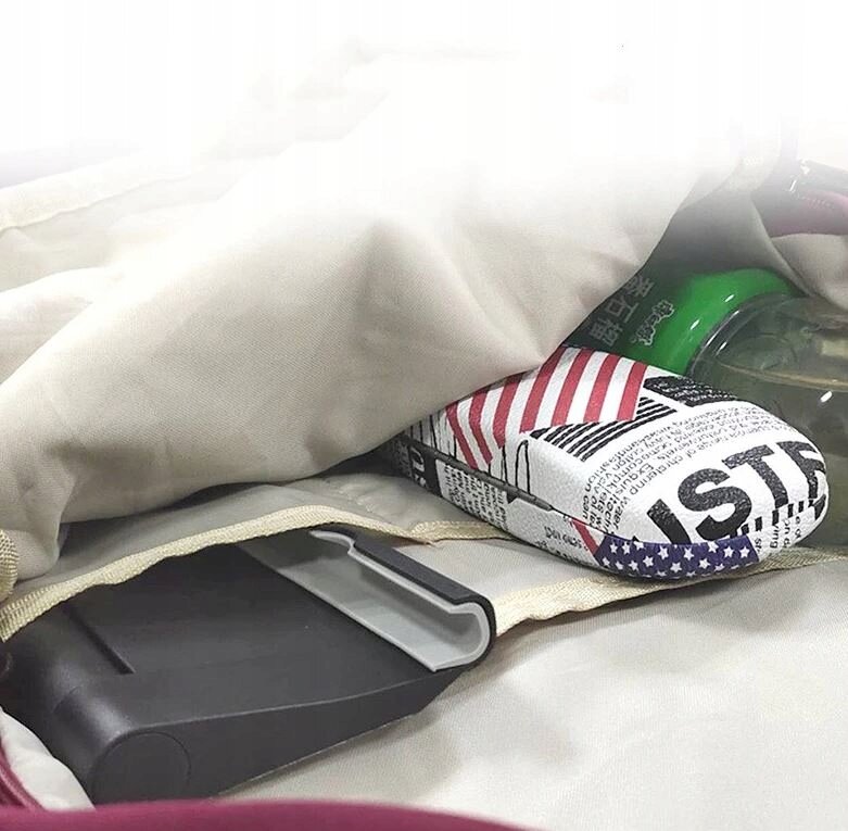 Na obrazie widoczny jest składany uchwyt na telefon w kolorze czarnym, leżący na jasnym materiale. Obok znajduje się przedmiot z wzorem przypominającym flagę USA oraz przezroczysta butelka z zieloną nakrętką.