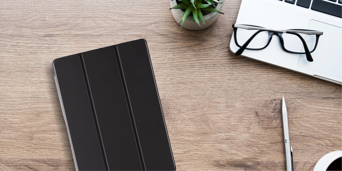 Na drewnianym biurku leży czarne etui na tablet, obok którego znajdują się okulary, laptop, długopis, filiżanka z napojem i mała roślina w doniczce.
