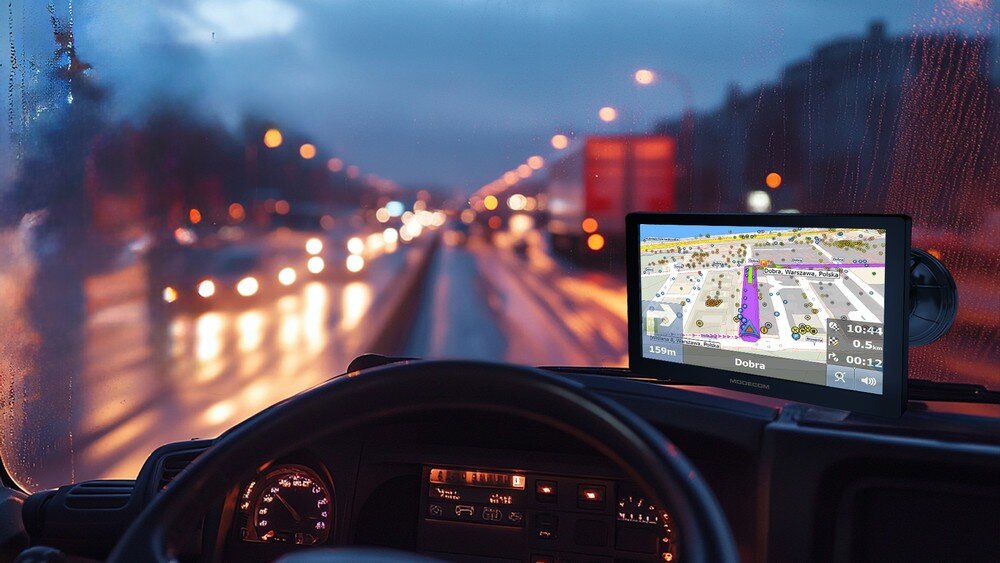 Widok z wnętrza pojazdu na drogę w deszczowy wieczór, z nawigacją GPS pokazującą mapę na ekranie. Na ekranie widoczny jest tekst: \'Dobra\'.
