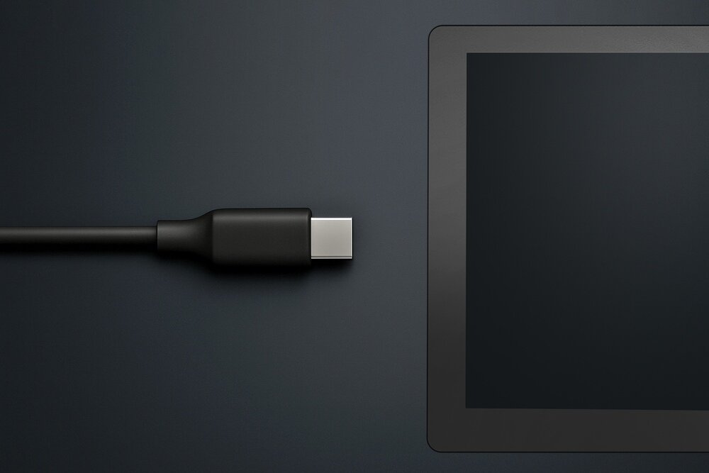 Obraz przedstawia czarny kabel USB-C obok czarnego urządzenia z ekranem, prawdopodobnie tabletu lub nawigacji. Tło jest ciemne i jednolite.