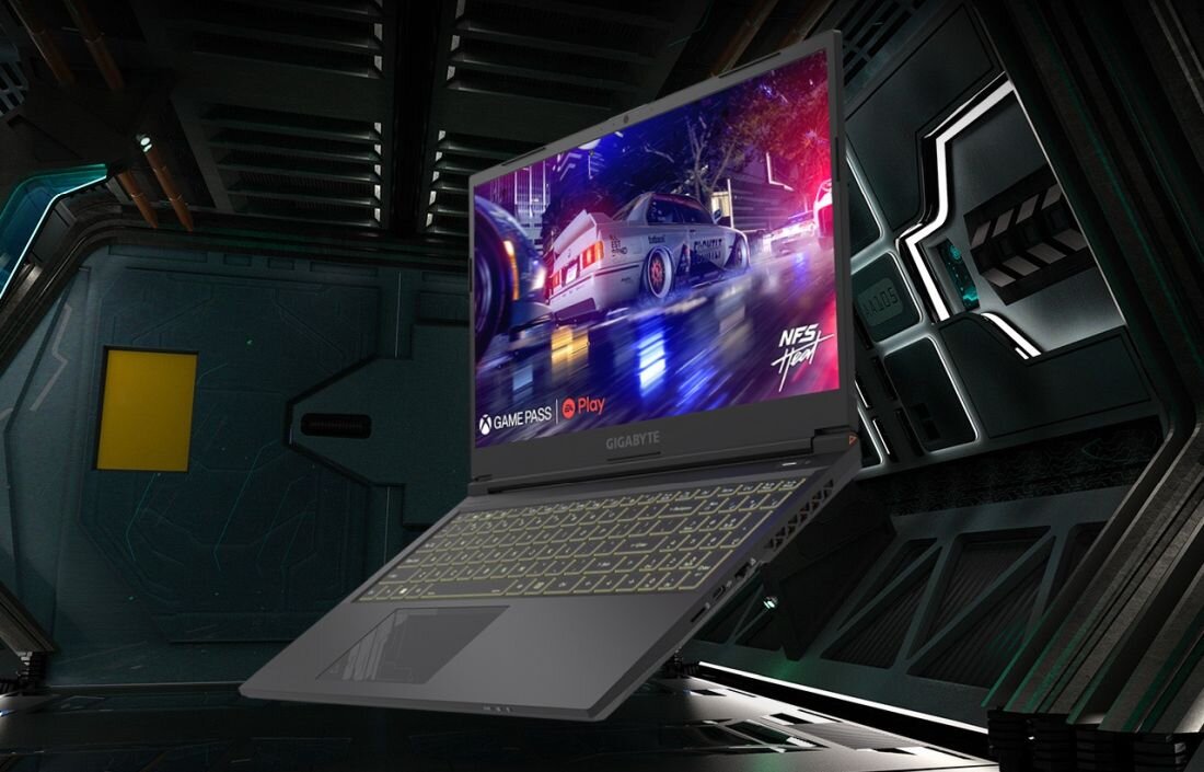 Laptop z wyświetlaczem pokazującym dynamiczną scenę z gry wyścigowej, z widocznymi logo \'GAME PASS\' i \'Play\'. Klawiatura jest podświetlona, a otoczenie ma futurystyczny wygląd.