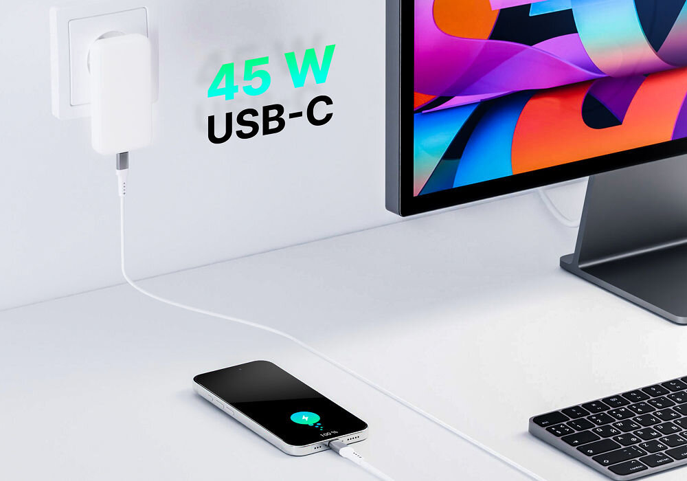 Biała ładowarka sieciowa podłączona do gniazdka, z kablem prowadzącym do telefonu leżącego na biurku. Obok widoczny jest monitor i klawiatura, a na ścianie napis \'45 W USB-C\'.