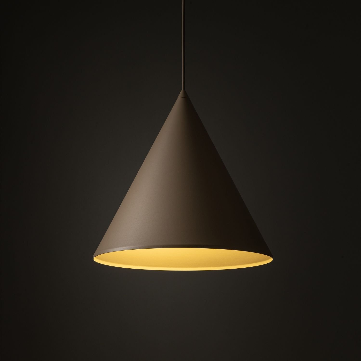 Na obrazie widoczna jest wisząca lampa w kształcie stożka, w kolorze beżowym, z jasnym wnętrzem. Lampa jest zawieszona na ciemnym tle.