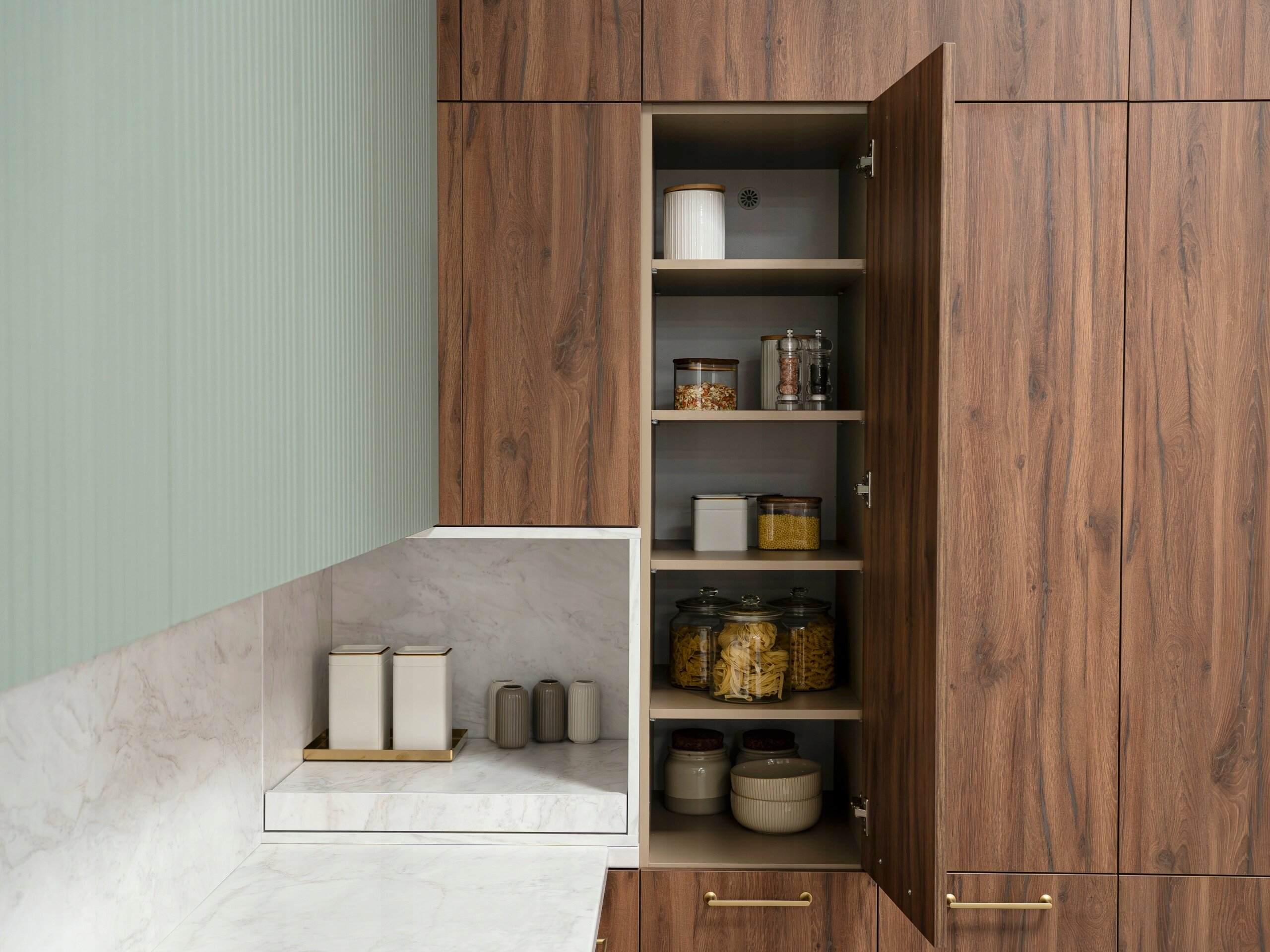 Zdjęcie przedstawia wnętrze kuchni z szafkami w kolorze drewna, z otwartą szafką ukazującą półki z pojemnikami na żywność. Na blacie znajduje się kilka pojemników w jasnych kolorach.
