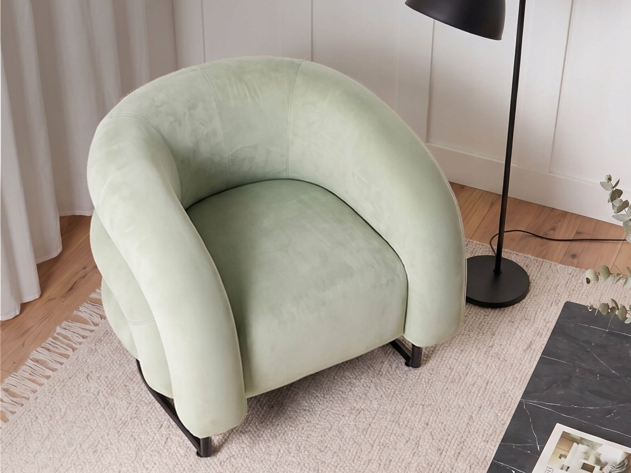 Miętowy, welurowy fotel wypoczynkowy o zaokrąglonym kształcie stoi na jasnym dywanie obok czarnej lampy stojącej. W tle widoczne są białe zasłony i drewniana podłoga.