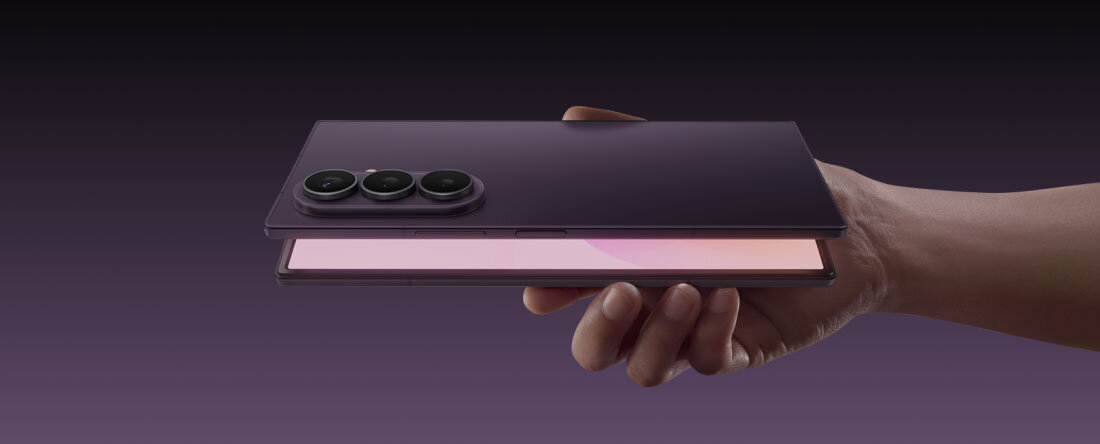 Galaxy Z Fold8 Ultra Osoba trzyma w dłoni składany smartfon w kolorze ciemnym, z trzema widocznymi obiektywami aparatu na tylnej części obudowy. Ekran urządzenia jest częściowo otwarty, ukazując jasną powierzchnię wewnętrzną.