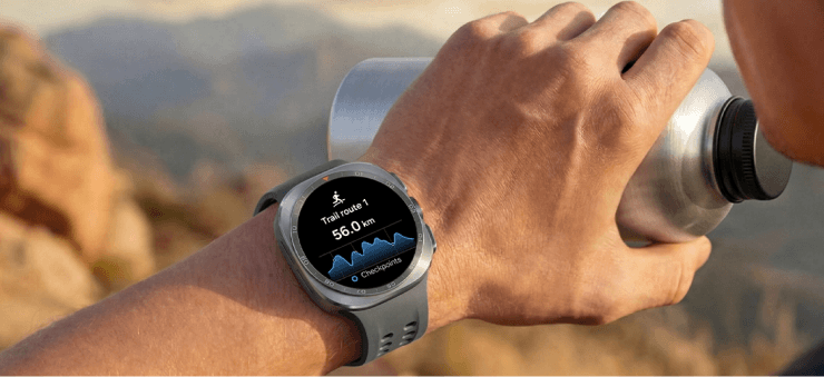 Dłoń ze smartwatchem podczas obserwacji górskiego krajobrazu. Osoba trzymająca metalowy bidon ma na nadgarstku smartwatch z wyświetlaczem pokazującym trasę biegu o długości 56.0 km. W tle widać rozmyty krajobraz górski.
