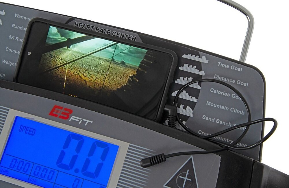 Panel sterowania bieżni z wyświetlaczem pokazującym prędkość 0.0 oraz telefonem umieszczonym w uchwycie. Widoczne są napisy: \'HEART RATE CENTER\', \'EB FIT\' oraz różne tryby treningowe takie jak \'Time Goal\', \'Distance Goal\'.