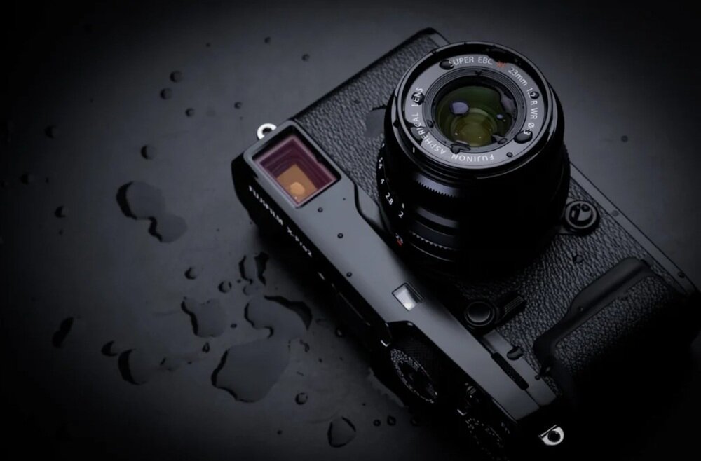 Czarny aparat fotograficzny z zamontowanym obiektywem FUJIFILM XF 23mm f/2.0 R WR, otoczony kroplami wody na ciemnym tle.
