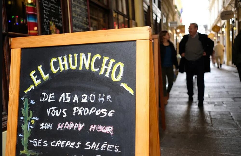 Tablica z napisem \'EL CHUNCHO DE 15 A 20 HR VOUS PROPOSE SON HAPPY HOUR SES CREPES SALEES\' stoi na ulicy, w tle widać przechodniów.