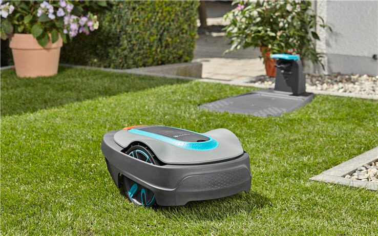 Na trawniku znajduje się robot koszący o nowoczesnym designie, z widocznymi niebieskimi i szarymi elementami. W tle widać stację dokującą oraz rośliny w doniczkach.