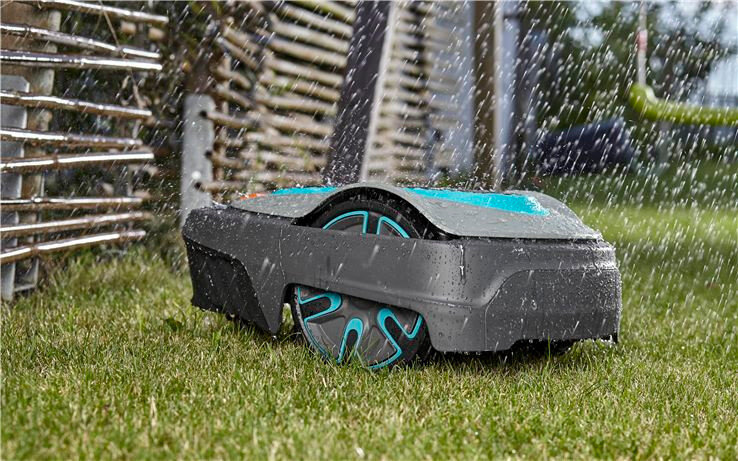 Robot koszący na trawie podczas deszczu, z widocznymi niebieskimi elementami na kołach i obudowie.