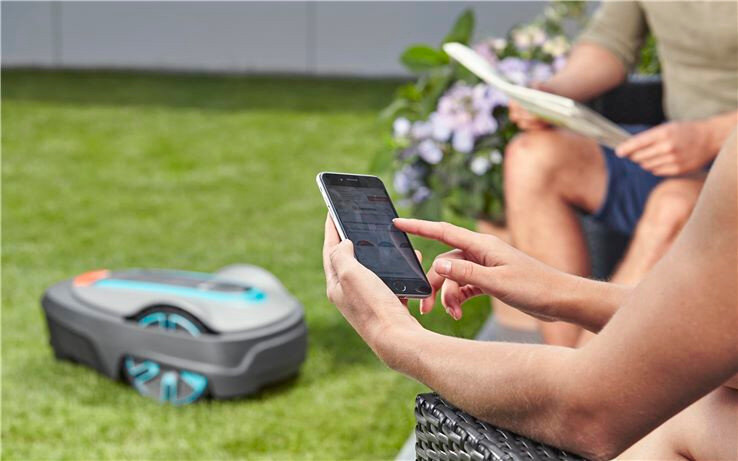 Osoba trzymająca smartfon siedzi na trawie, a w tle widać robot koszący na trawniku. Obok znajduje się druga osoba z gazetą, a w tle są kwiaty w donicy.