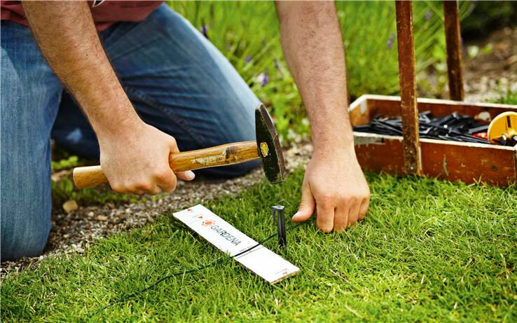 Osoba wbija czarny kołek w trawnik za pomocą młotka. Obok znajduje się biały pasek z logo \'GARDENA\'.