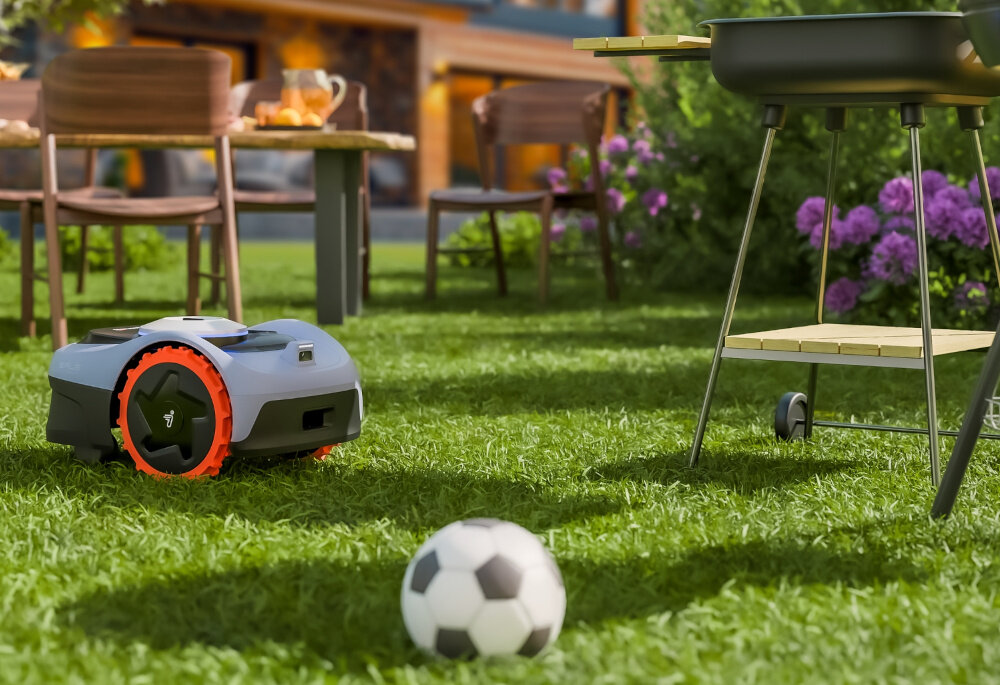 Na trawniku znajduje się robot koszący z pomarańczowymi kołami oraz piłka do piłki nożnej. W tle widoczny jest stół z krzesłami i grill, a także kwitnące krzewy.