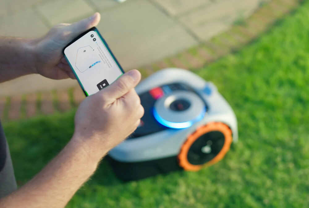 Osoba trzyma smartfon z aplikacją pokazującą mapę trawnika, a w tle widoczny jest robot koszący na trawie.