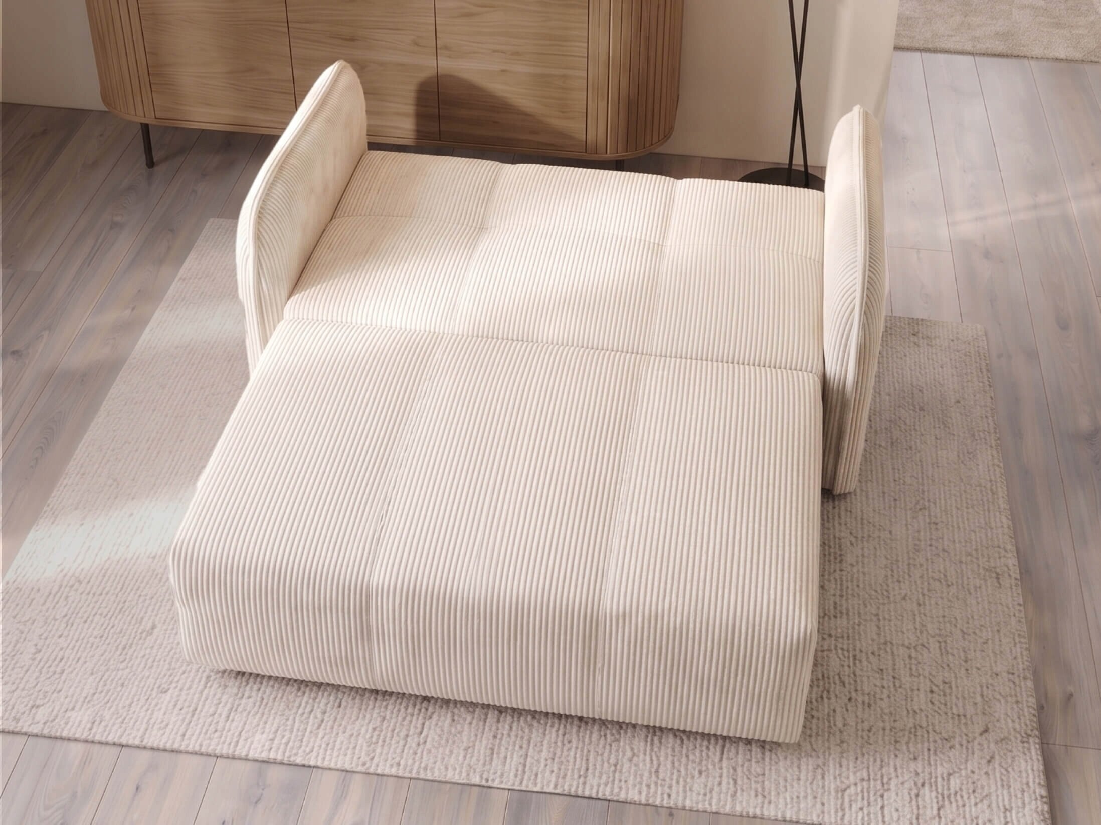 Rozkładana sofa w kremowym kolorze z widocznymi pionowymi przeszyciami, ustawiona na jasnym dywanie w pomieszczeniu z drewnianą podłogą.