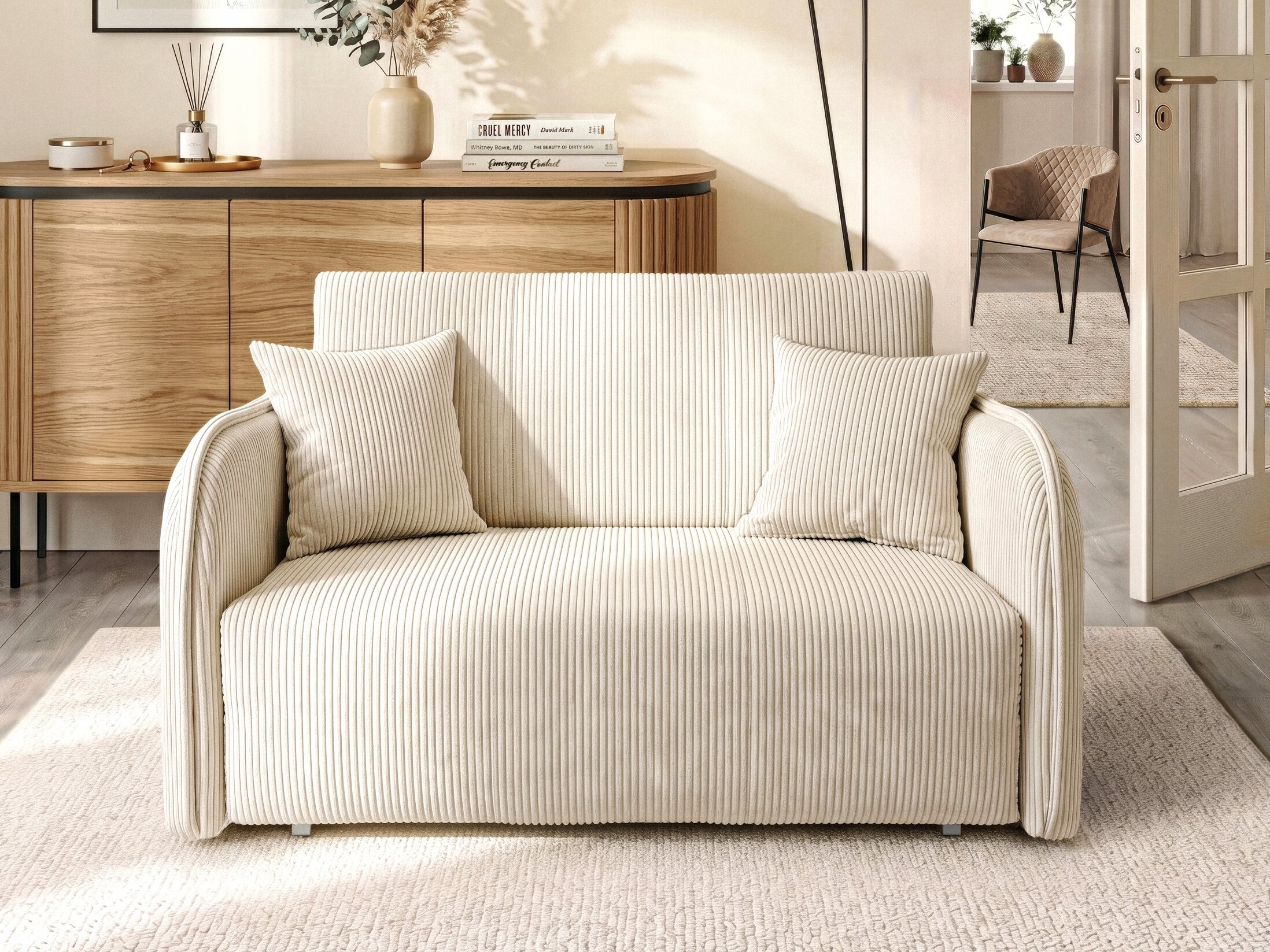 Kremowa sofa rozkładana z dwoma poduszkami stoi na jasnym dywanie w eleganckim wnętrzu. W tle znajduje się drewniana komoda z dekoracjami i książkami.
