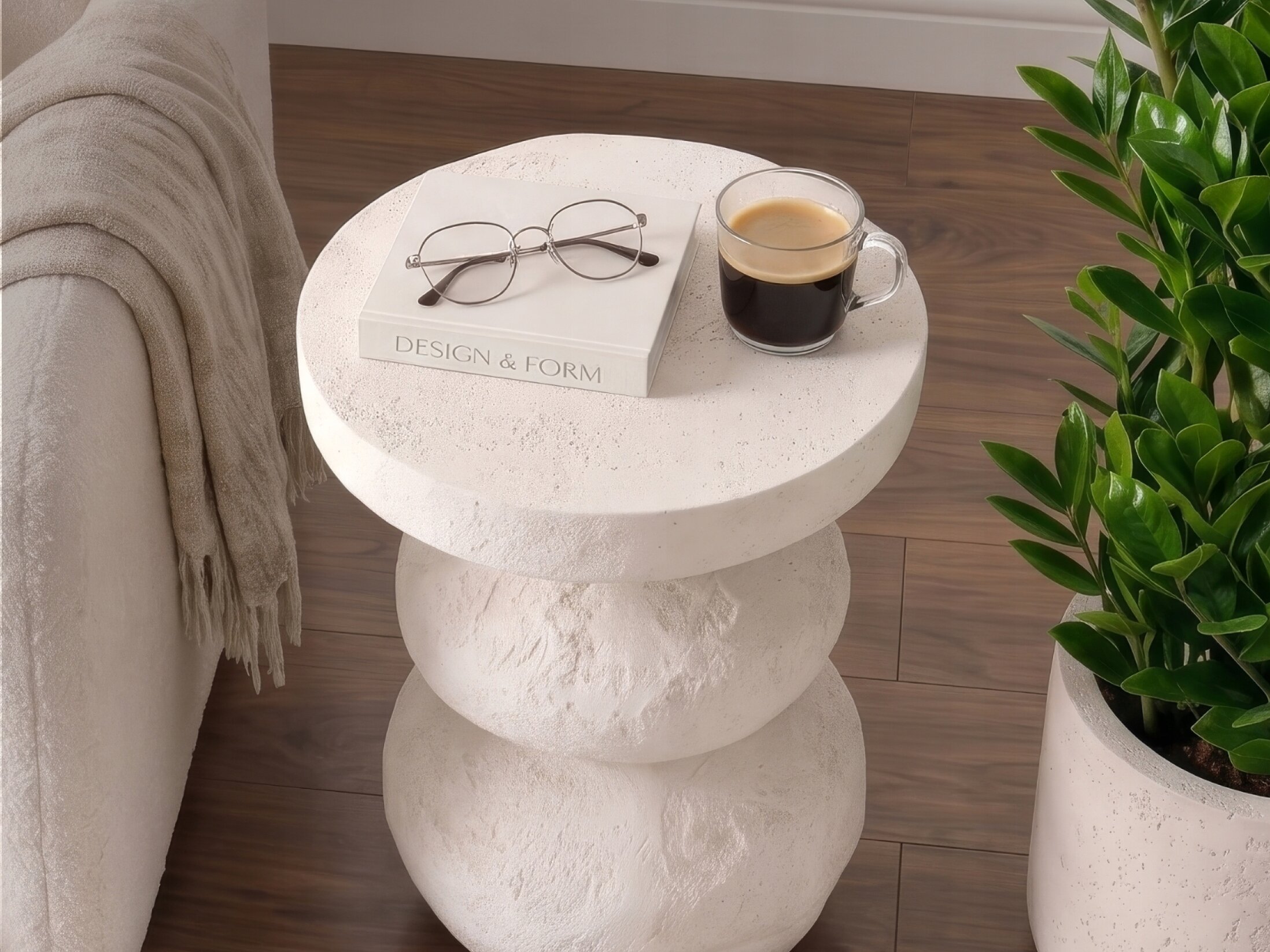 Na okrągłym kremowym stoliku kawowym znajduje się książka z napisem \'DESIGN & FORM\', okulary oraz filiżanka kawy. Obok stolika stoi roślina w doniczce, a w tle widoczna jest część sofy z kocem.