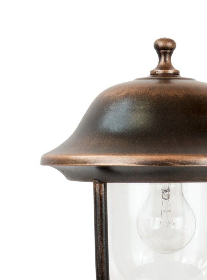 Lampa zewnętrzna stojąca w kolorze patyny miedzi z widoczną żarówką wewnątrz szklanego klosza. Górna część lampy ma zaokrąglony kształt z ozdobnym zakończeniem na szczycie.