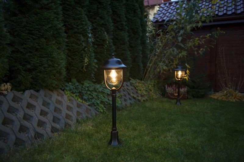 Na trawniku w ogrodzie stoją dwie zewnętrzne lampy o klasycznym wyglądzie, emitujące ciepłe światło. W tle widoczne są wysokie krzewy i fragment budynku z dachówką.